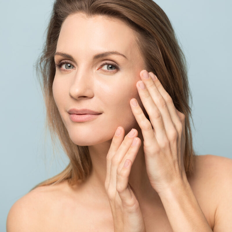 Beautiful millennial Caucasian naked woman isolated on blue studio background touch soft healthy face skin pleased with beauty procedures, pretty blonde female satisfied with clinic facial injection